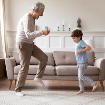 Full length side view overjoyed older senior grandfather dancing with excited little grandson in living room. Happy different generations family having fun, enjoying favorite music together at home.